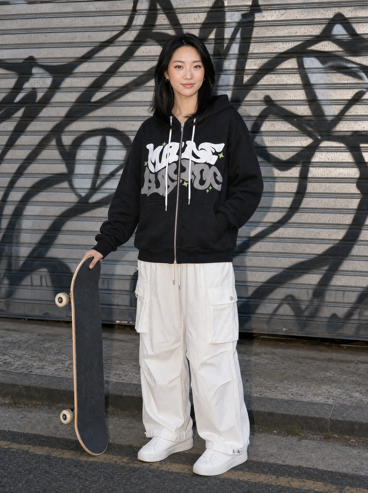 Photo lifestyle | Femme devant un rideau métallique tagué, skate à la main, sweat zippé noir à lettrage graffiti et cargo blanc | Variante : Noir