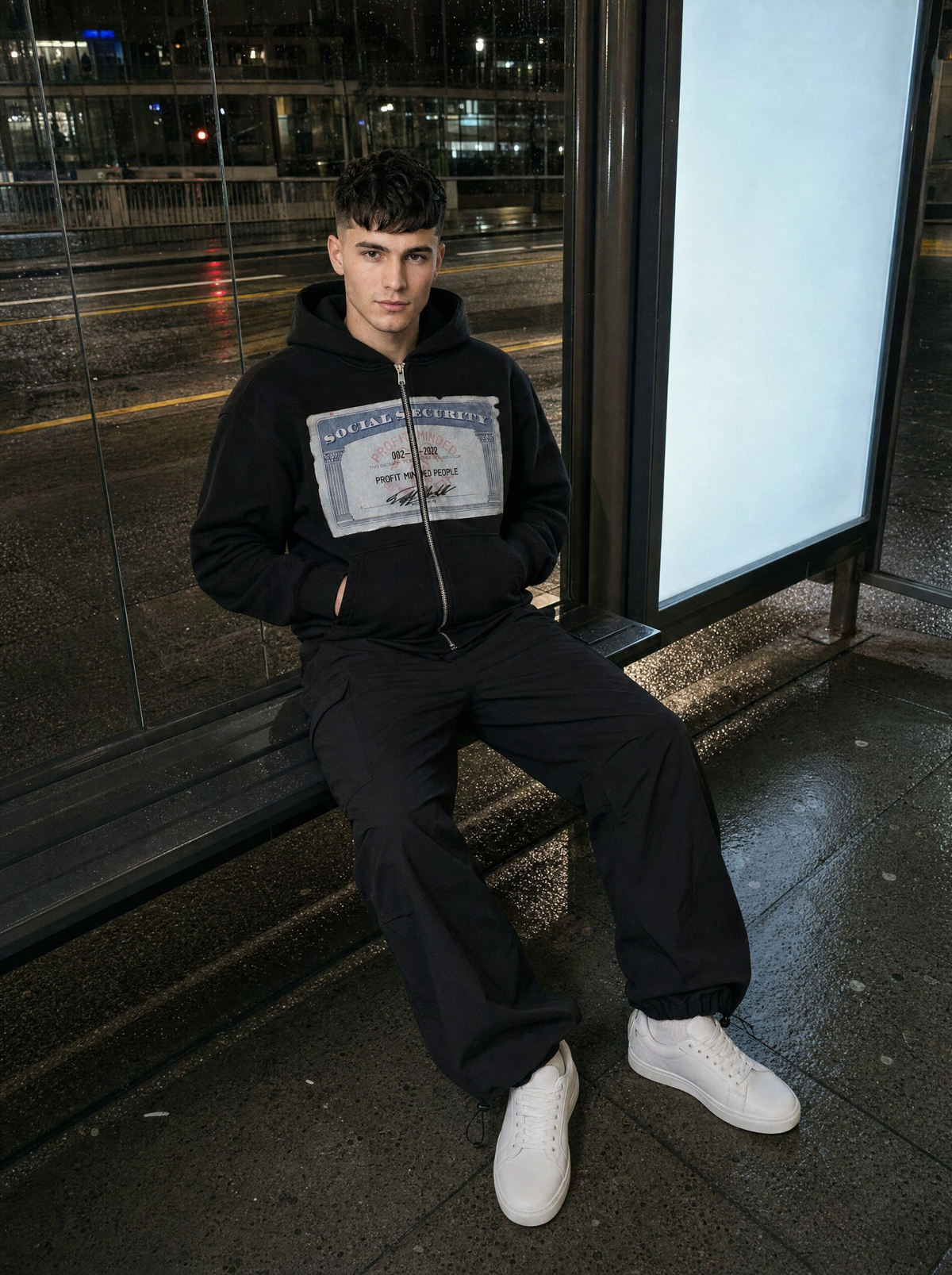Photo lifestyle | Hoodie zippé noir à imprimé carte rétro porté par un homme assis sous un abribus de nuit, avec un pantalon parachute noir | Variante : Noir