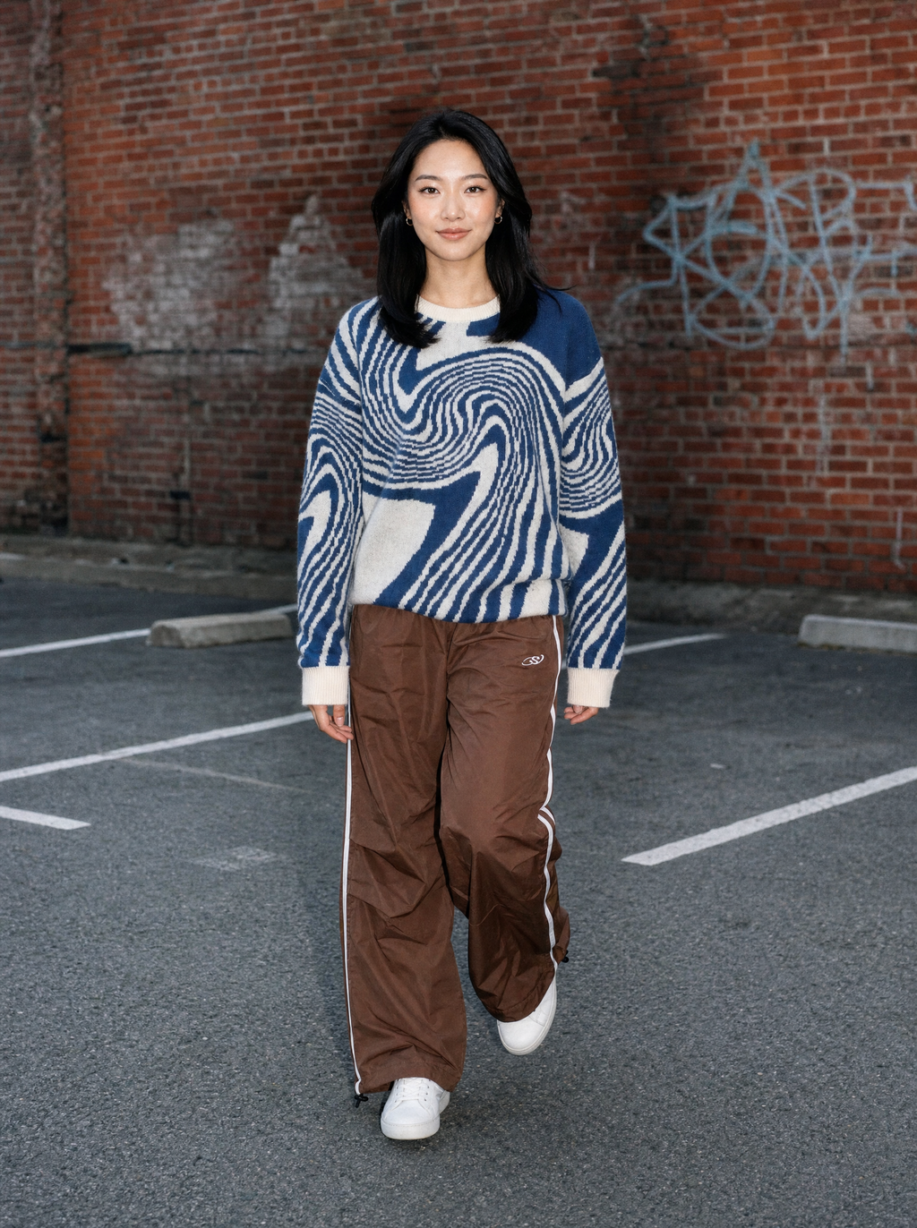 Photo lifestyle | Jogging large marron en nylon à bandes latérales blanches porté par une femme dans un parking en brique, tenue streetwear | Variante : Marron