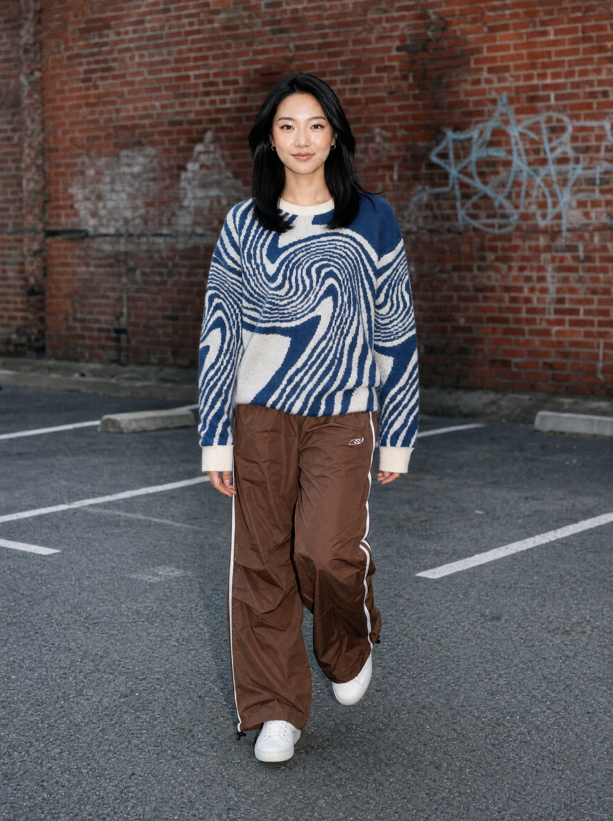 Photo lifestyle | Jogging large marron en nylon à bandes latérales blanches porté par une femme dans un parking en brique, tenue streetwear | Variante : Marron