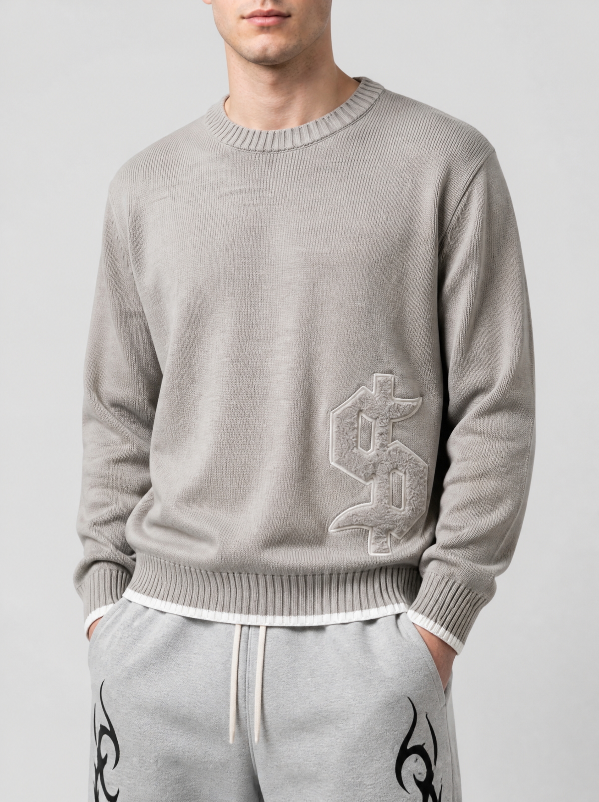 Photo détail | Gros plan sur le signe dollar gothique en relief contouré de blanc sur le bas du pull en maille taupe