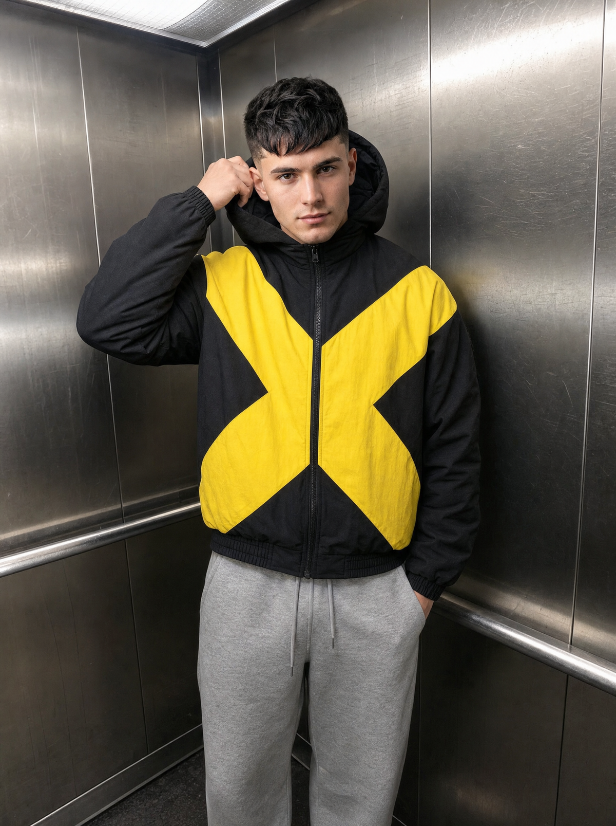 Photo lifestyle | Homme dans un ascenseur en métal, bomber noir rembourré à grand X jaune vif, capuche et jogging gris