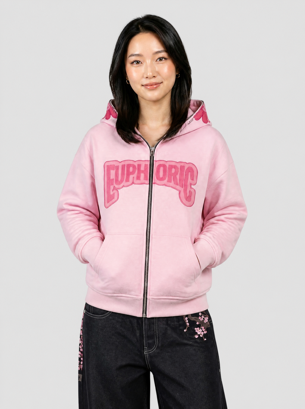Autre vue | Femme de face en veste zippée rose à capuche fleur souriante et inscription Euphoric, portée avec un jean noir large