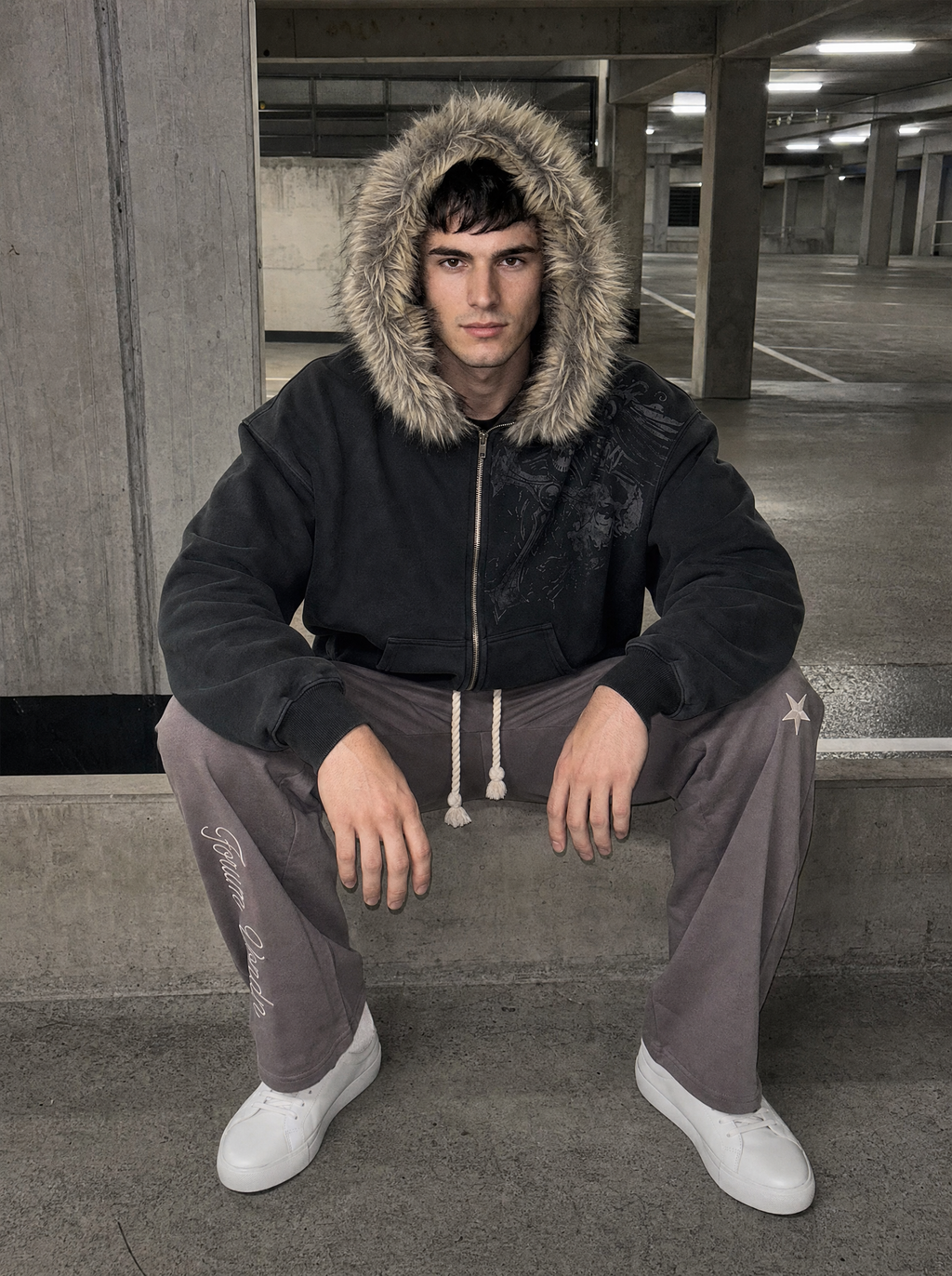 Photo lifestyle | Homme accroupi dans un parking souterrain, veste zippée noire délavée à capuche en fausse fourrure et jogging gris | Variante : Noir