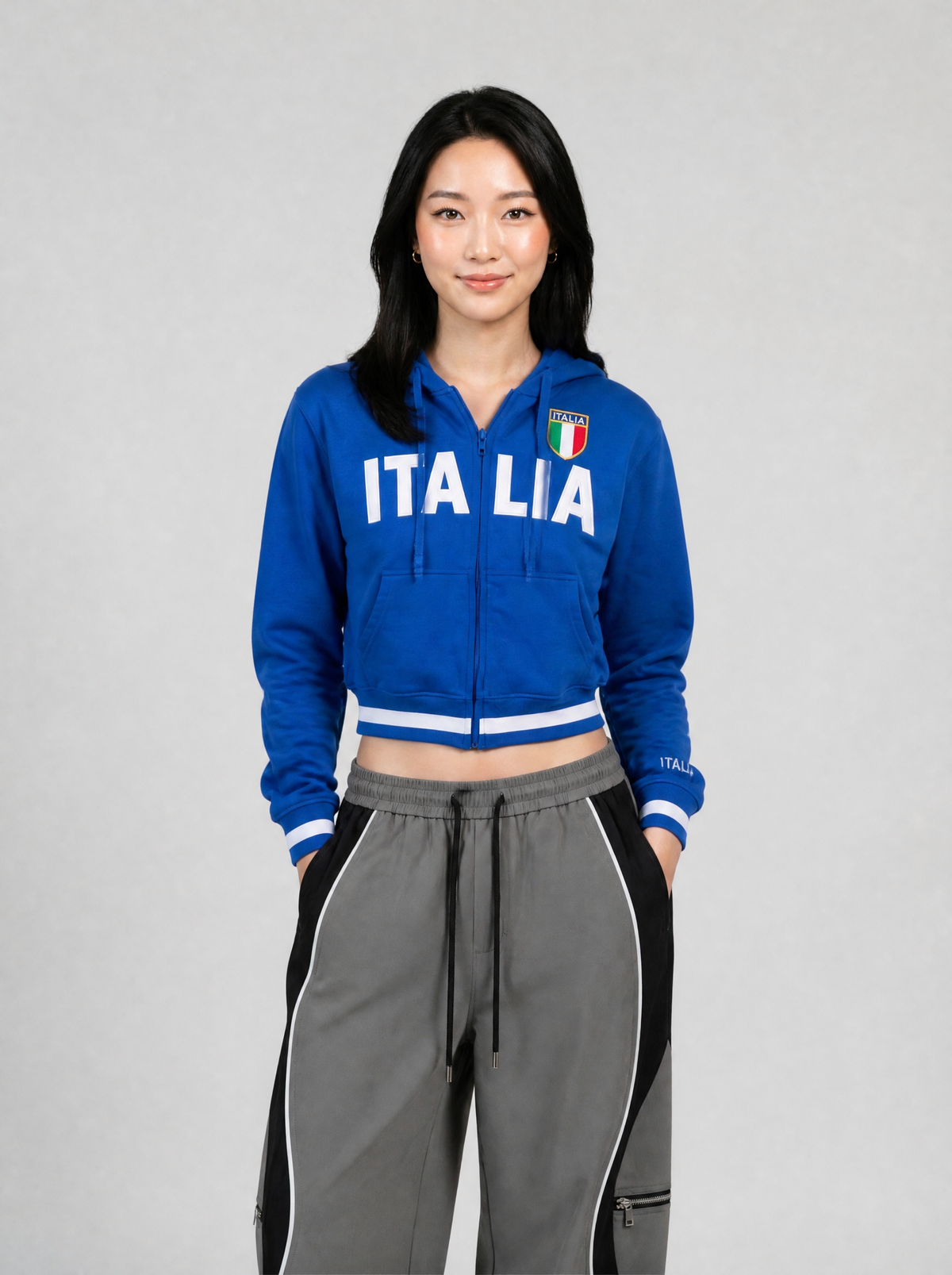 Autre vue | Femme de face en veste zippée bleu roi courte avec lettrage Italia blanc et bords rayés, avec un pantalon large gris