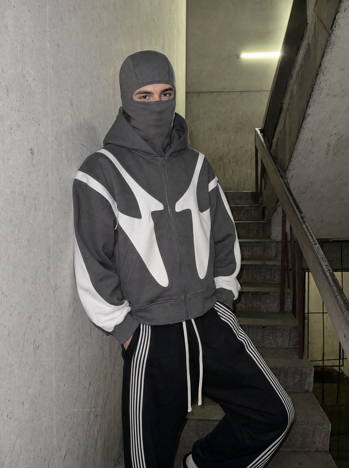 Photo lifestyle | Homme adossé au mur d'une cage d'escalier en béton, veste zippée grise à cagoule relevée et panneaux blancs, jogging noir | Variante : Gris