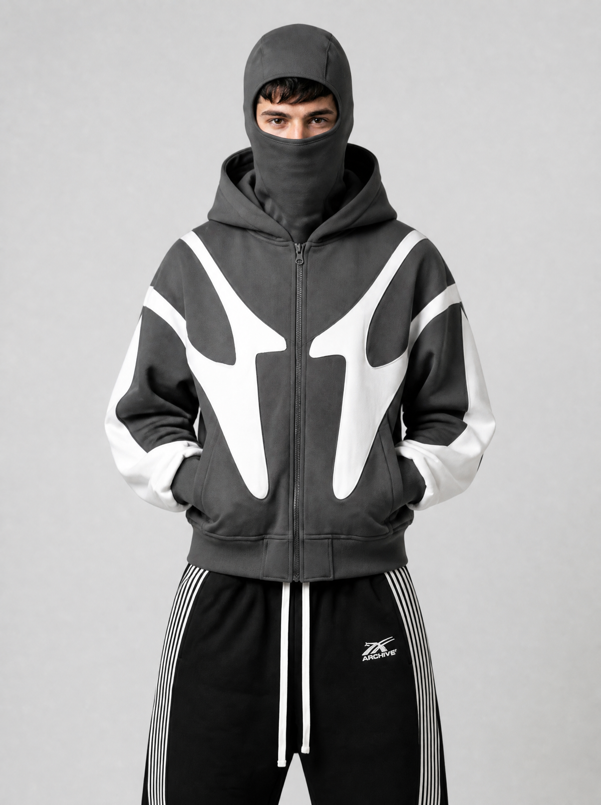 Autre vue | Homme de face en veste zippée grise cagoule relevée, panneaux blancs en forme de crocs, avec un jogging large noir | Variante : Gris