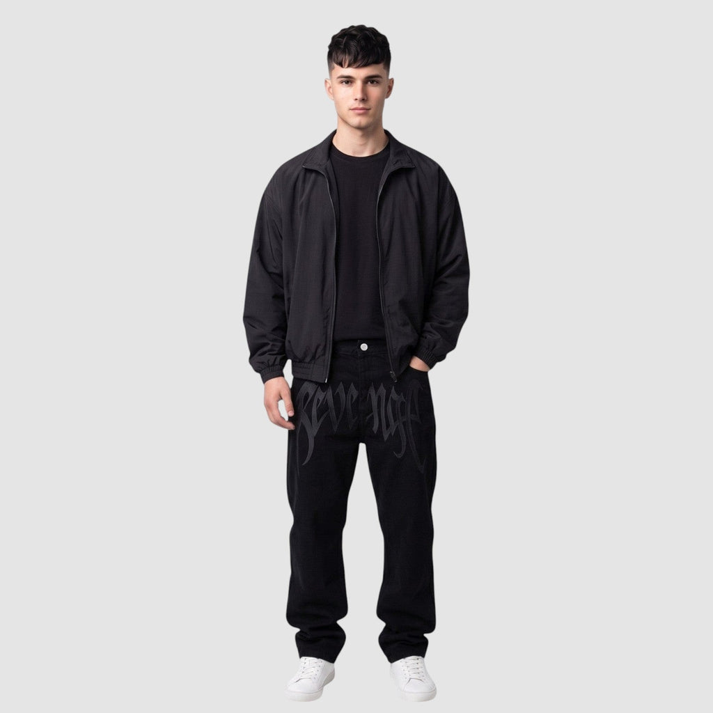 Y2K Avenue black Y2K hip-hop jeans with gothic lettering on thighs, worn by a model with a black jacket and sneakers