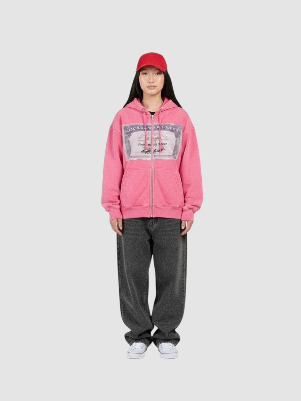 Y2K Avenue pink zip-up hoodie with a graphic card print on the chest, worn by a model with a red cap and grey jeans.
